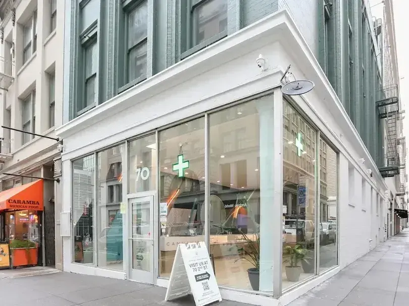 Corner storefront of a cannabis dispensary with glass windows and green cross sign